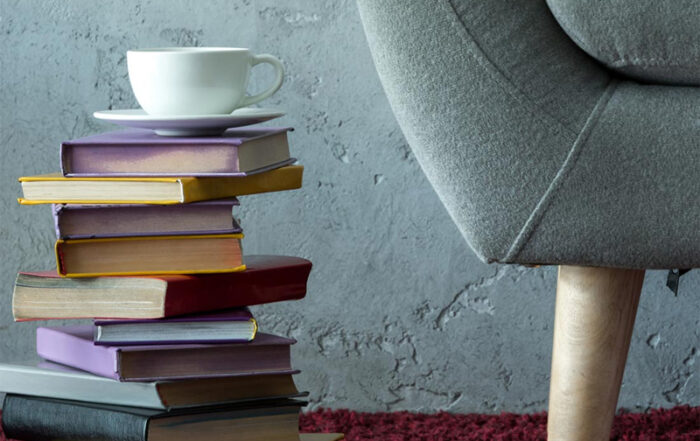 Das Foto zeigt einen hohen Stapel unterschiedlich großer Bücher, auf dem eine weiße Tasse mit Untertasse abgestellt ist. Daneben steht ein gepolsterter Sessel auf einem dunkelroten Teppich vor einer grau strukturierten Wand.