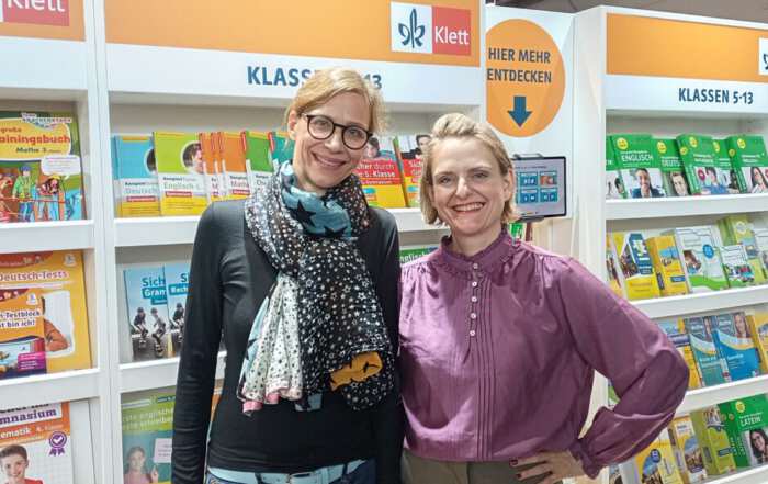 Das Foto zeigt zwei Frauen vor einem Messestand mit Regalen voller Schul- und Unterrichtsbücher. Im Hintergrund sind zahlreiche Buchcover, Verlagslogos und Hinweisschilder zu sehen, darunter das Klett-Logo und die Aufschrift „Hier mehr entdecken“.