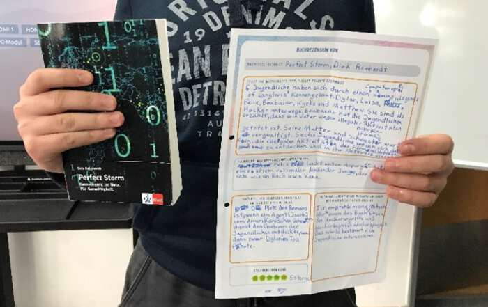 Das Foto zeigt eine Person, die ein Buch mit dem Titel „Perfect Storm“ sowie ein handschriftlich ausgefülltes Arbeitsblatt zur Buchrezension in die Kamera hält. Im Hintergrund sind eine Tafel oder ein Bildschirm und ein Klassenraum zu erkennen.