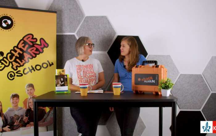 Das Bild zeigt zwei Personen an einem Tisch in einem Studio mit sechseckigen Akustikelementen an der Wand. Auf dem Tisch stehen Tassen, ein Buchaufsteller und ein orangefarbener Koffer mit der Aufschrift „BÜCHER ALARM“; links ist ein Banner mit dem Schriftzug „Bücheralarm @ School“ zu sehen.